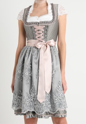 Femme portant une robe traditionnelle grise avec broderies florales, manches en dentelle, laçage rose sur le corsage et un grand nœud rose à la taille.