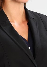 Černý oblekový blejzr s klopovým límcem, texturovanou tkaninou a detailním šitím. Nositý přes námořnický top, představující profesionální vzhled.