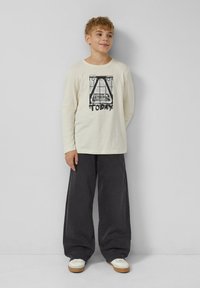 Kremowa bawełniana koszulka z długim rękawem z czarnym nadrukiem graficznym i napisem „TODAY” na przodzie, zestawiona z szerokimi szarymi spodniami dżinsowymi i białymi tenisówkami.