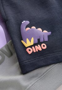 Tissu bleu marine avec une applique de dinosaure violet et le texte orange "DINO", posé sur un matériau gris. Présente des textures en relief et des couleurs contrastées.