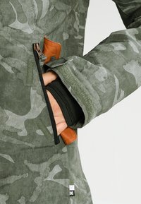 Grön kamouflagejacka med ett mjukt innerfoder, dragkedjefickor och detaljer i brunt läder. Har ett strukturerat, slitstarkt yttertyg.