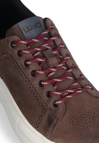 Brązowe zamszowe sneakersy z białą podeszwą akcentującą, ozdobione burgundowymi i białymi sznurówkami w paski, perforacjami oraz etykietą z logo na języku.