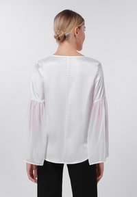 Blouse en satin blanc avec des manches longues et bouffantes semi-transparentes ; encolure ronde ; texture lisse et subtil éclat ; conçue avec une coupe droite.