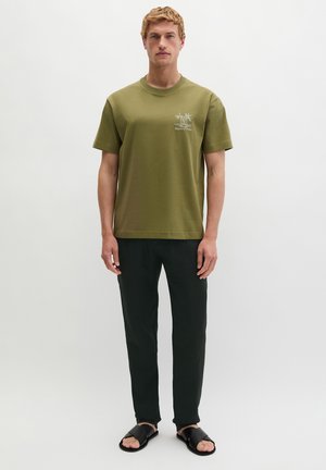 Homme portant un t-shirt vert olive à manches courtes avec un motif de palmiers, pantalon noir et sandales noires, debout devant un fond gris uni.