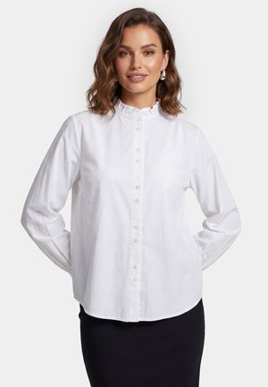 Vrouw met schouderlang bruin haar, zilveren oorringen, een witte blouse met knopen en een gerimpelde kraag, en een zwarte rok, staand met de handen achter.