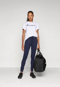 Bela majica s kratkimi rokavi in mornarsko modrim logotipom, kombinirana z mornarsko modrimi trenirkami z zračnim trakom, črnimi supergami in črno torbo za šport.