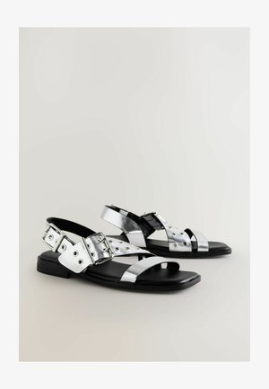Zilverkleurige metallic sandalen met platte zwarte zolen, voorzien van kruisstappen en oogjesdetails. Verstelbare gesp-sluiting op de enkelriem.