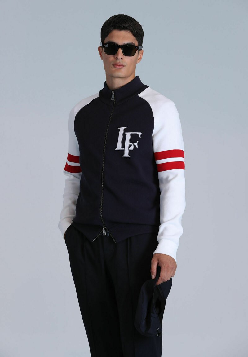 Sportliche Zip-Jacke in Marineblau und Weiß mit roten Streifenakzenten, mit einem Logo auf der Brust und kontrastierenden Ärmeln. Glatte Textur.