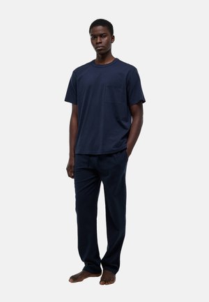 Jeune homme debout pieds nus, portant une chemise à manches courtes bleu marine avec une poche sur la poitrine et un pantalon assorti bleu marine texturé.