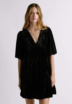 Vestido de terciopelo negro con un escote en V profundo, mangas cortas y una cintura fruncida. La tela tiene una textura acanalada y cae de manera suelta.
