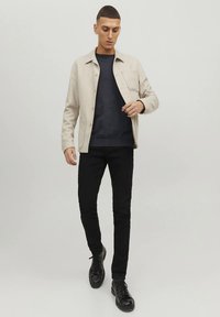 Beige overhemdjack gedragen over een donkerblauwe trui en zwarte skinny jeans, gecombineerd met zwarte veterlaarzen. Overal gladde texturen.