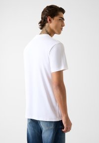 Biała koszulka z krótkim rękawem i okrągłym dekoltem, wykonana z gładkiego materiału. Modelka nosi ją wpuszczoną w luźne niebieskie dżinsy.