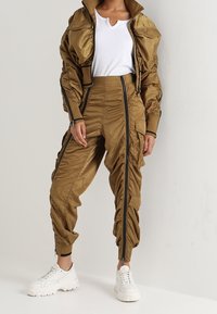 Gouden satijnen trainingspak met een hoge kraagjack en gekreukte broek, voorzien van zwarte ritsen, gecombineerd met witte chunky sneakers.