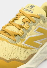 Gul mesh-sneaker med vadderad tunga, framträdande "N"-logotyp, randiga snören och texturerad detalj på ovandelen.