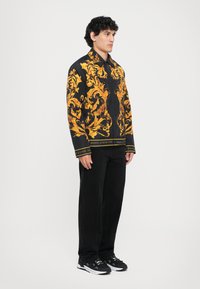 Bărbat în picioare, îmbrăcat cu o jachetă neagră cu modele baroce aurii intricate și mari, pantaloni negri și adidași alb-negru, întors spre dreapta.