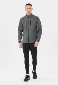 Graue Jacke mit hohem Kragen, Reißverschlusstasche auf der Brust und langen Ärmeln, kombiniert mit schwarzen Leggings und schwarzen Turnschuhen. Glatte Textur.
