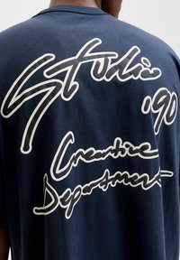 Marineblå t-skjorte med stor hvit og teksturert svart skrift på baksiden, som sier "Studio '90 Creative Department."