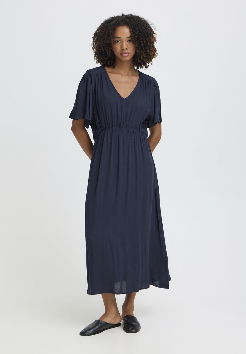 Femme aux cheveux bouclés portant une robe midi bleu marine à col en V avec de courtes manches volantées et des chaussures noires sans lacets, debout devant un fond blanc.