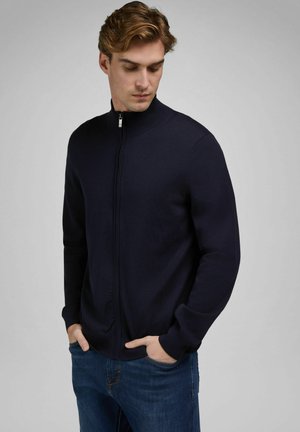 Junger Mann mit den Händen in den Taschen, trägt einen dunkelblauen, Reißverschluss-Pullover und blaue Jeans, schaut nach unten links vor einem einfarbigen grauen Hintergrund.