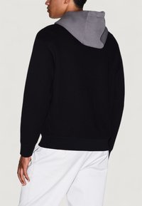 Hombre vistiendo una sudadera negra con capucha gris, combinado con pantalones blancos, de pie de lado frente a un fondo sencillo.