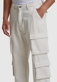 Pantalones cargo crema con múltiples bolsillos laterales, cremallera y cierre de botón. Fabricados con un material duradero y acabado texturizado.