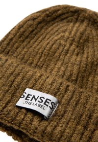 Braune gestrickte Mütze mit einem umgeschlagenen Rand und einem weißen Stoffetikett, auf dem "SENSES THE LABEL" auf der Manschette genäht ist.
