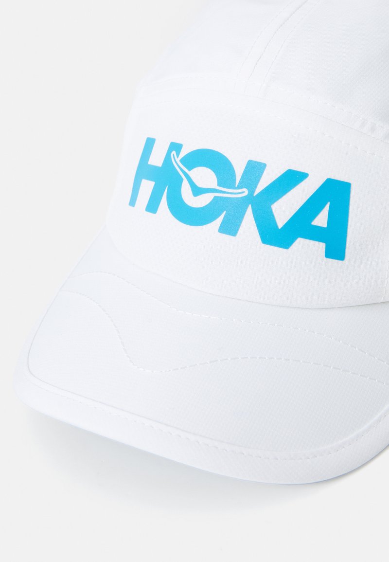 White cap made of lightweight material featuring a prominent blue "HOKA" logo. Smooth texture with curved stitching details on the front.