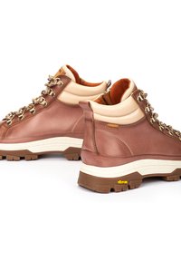 Botas de senderismo de cuero marrón con detalles beige, ojales metálicos y suela blanca Vibram. Diseño de caña alta para soporte y durabilidad.