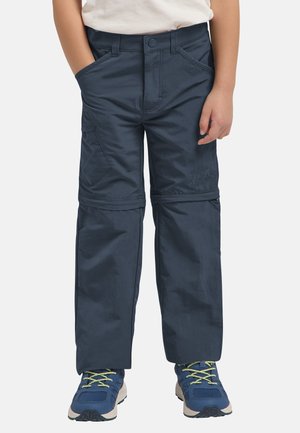 Kind trägt marineblaue Wanderhose mit Reißverschluss zum Abzippen und blaue Turnschuhe mit grünen Schnürsenkeln, steht mit Händen in den Taschen.