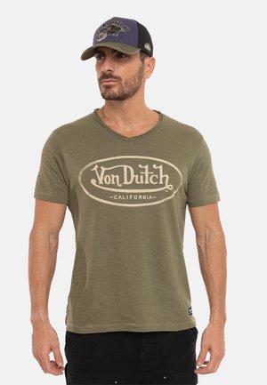 Homme portant un t-shirt vert olive Von Dutch et une casquette noire avec le logo Von Dutch, debout devant un fond clair uni.