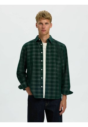 Jeune homme aux cheveux blonds courts portant une chemise à carreaux vert foncé boutonnée par-dessus un t-shirt blanc et un pantalon foncé, debout devant un fond uni.