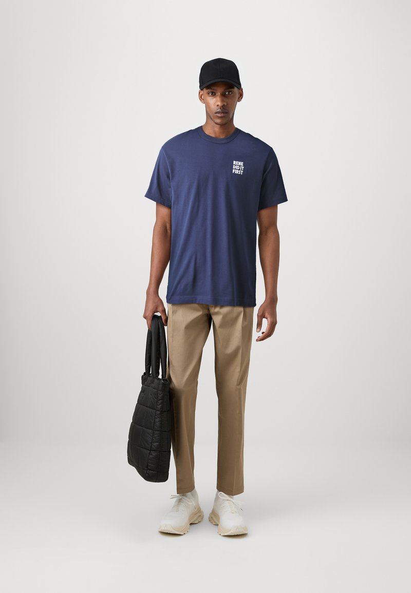 Camiseta de algodón azul marino con texto en el lado izquierdo del pecho, pantalones beiges, zapatillas blancas y un bolso de mano negro acolchado.