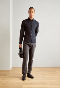 Camisa negra de botones, pantalones grises, mocasines negros, sosteniendo un pequeño bolso negro. Camisa de textil suave, diseño ajustado y estilo casual.