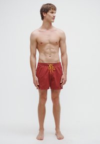 Punased ujumispüksid kergest kangast, millel on kontrastset kollast värvi pael. Disain on lihtne, pikkusega kuni reie keskmise osa.