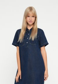 Denim shirt dress in dark blue with a collared neck, short sleeves, and two chest pockets. Features button-down front and loose fit.