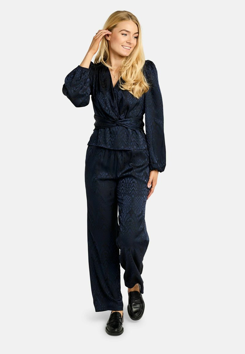 Tumesinise kaheosaline komplekt, millel on tekstureeritud pigemumplu top ja laiade säärtega püksid. Pehme, läikiv kangas peene mustriga, kombineeritud mustade kingadega.