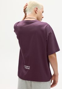 Lilla bomulds t-shirt med afslappet pasform, med korte ærmer og hvid broderet tekst på den nederste del af ryggen. Glat, enkel design.