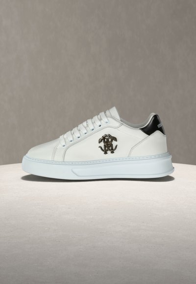 Λευκό sneaker χαμηλού παπουτσιού με μαύρη ταμπέλα στη φτέρνα, διακοσμητικό μεταλλικό λογότυπο στο πλάι, λευκά κορδόνια και παχιά γαλάζια σόλα σε μπεζ επιφάνεια.