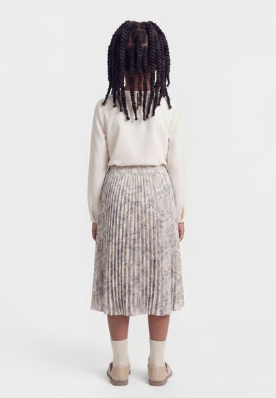 Παιδί με μακριά στριφογυριστά κοτσίδια, που φοράει κρεμ πουλόβερ, φούστα με πλισέ σχέδιο, κάλτσες κρεμ και μπεζ παπούτσια, στέκεται με την πλάτη γυρισμένη σε λευκό φόντο.