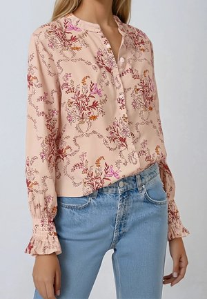 Kvinde iført en sart lyserød blomsterbluse med flæsemanchetter og lyseblå jeans med højtaljet pasform mod en ensfarvet baggrund.