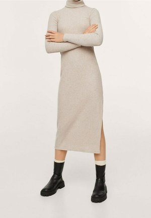 Vestido de punto beige con cuello vuelto, mangas largas y abertura lateral, combinado con botas negras hasta el tobillo y calcetines cremosos contrastantes. Textura suave.