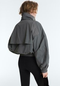 Personne aux cheveux courts portant une veste ample gris foncé à col montant et des leggings noirs, tenant une sangle ou un sac noir.