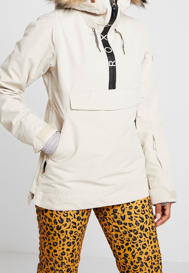Beige Kapuzenjacke mit schwarzem Reißverschluss und Logo, großer Fronttasche und Kunstfellbesatz. Kombiniert mit gelben Leopardenmuster-Hosen.