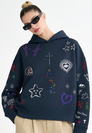 Femme portant un sweat à capuche bleu marine avec des gribouillis colorés incluant des étoiles, des cœurs, des oiseaux, un crâne et des motifs célestes, assorti à des lunettes de soleil noires.