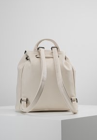 Mochila de cuero color crema con forma estructurada, cierre de cordón y correas ajustables. Presenta acentos de hardware plateado.