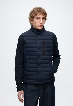 Giovane uomo con capelli ricci che indossa un gilet trapuntato blu navy sopra una maglia nera a maniche lunghe e jeans scuri, in piedi davanti a uno sfondo chiaro e semplice.
