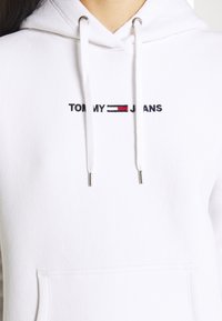 Sudadera blanca de tela suave con un logotipo bordado de "TOMMY JEANS", con un diseño en rojo y azul marino, y ojetes de metal plateado en la capucha.