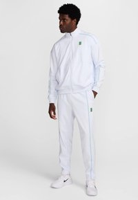 Bílá tepláková souprava se světle modrými pruhy, obsahující bundu na zip a odpovídající kalhoty. Na bundě a kalhotách je logo.