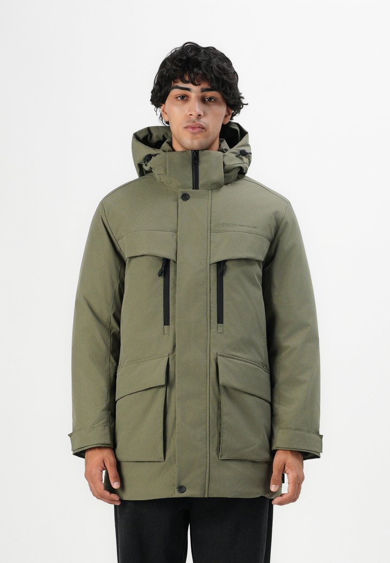 Mann in einem olivgrünen Winterparka mit Kapuze und mehreren mit Reißverschlüssen und Knöpfen versehenen Taschen, der vor einem schlichten weißen Hintergrund steht.