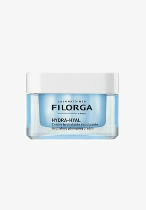 Genomskinlig glasburk med ljusblå botten och vit lock. Etiketten visar produktnamnet "HYDRA-HYAL" och "Återfuktande uppplumpningskräm" i svart text.